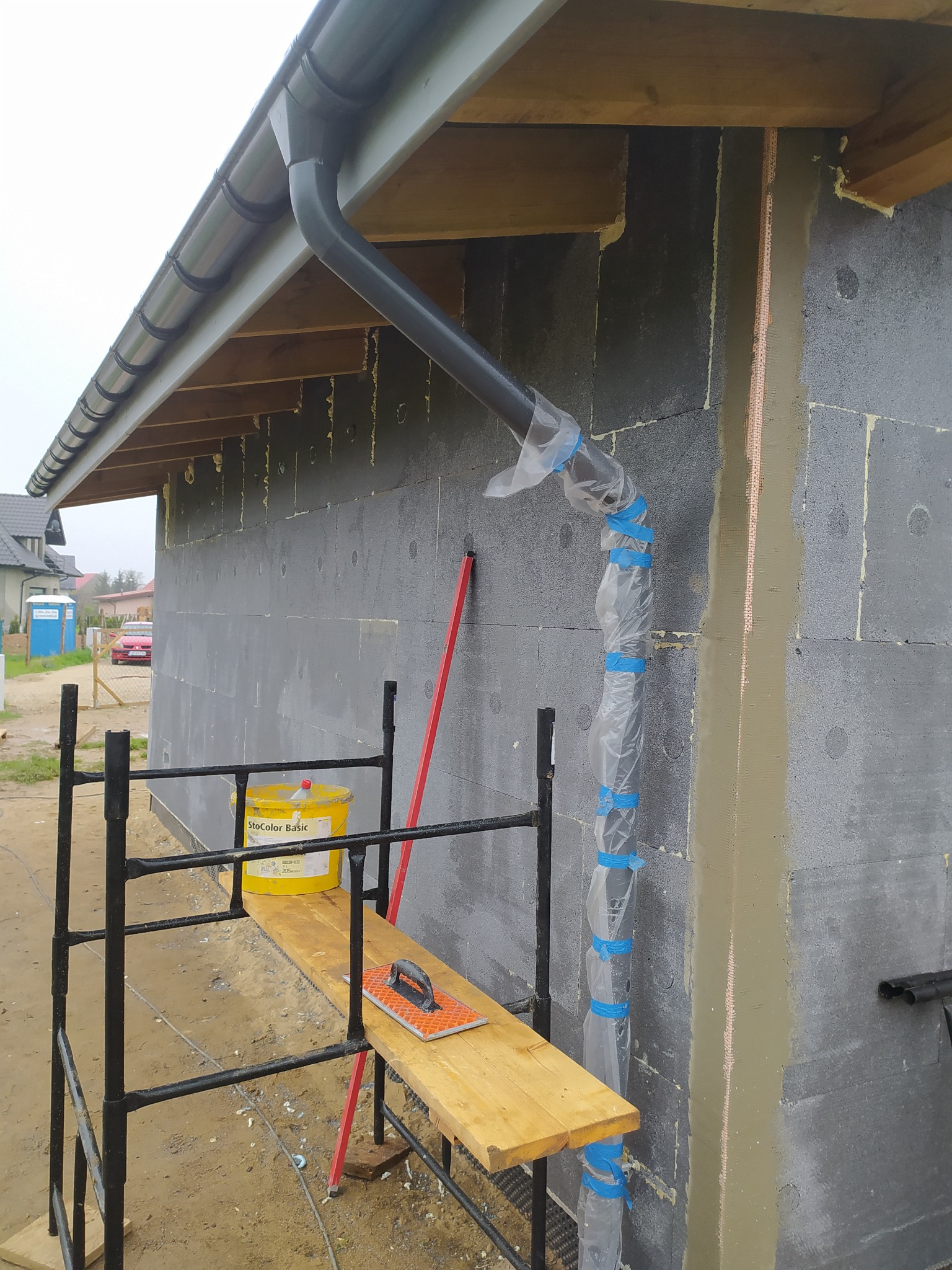 Ocieplanie ściany budynku z szarego bloczka styropianem, widoczny fragment rusztowania z wiadrem farby StoColor Basic i pacą do gładzi, rura spustowa owinięta folią zabezpieczającą.