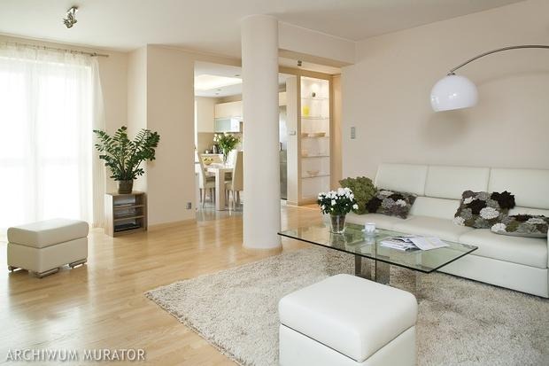 Jasne, przestronne wnętrze salonu z widokiem na jadalnię i kuchnię, z białymi meblami, szklanym stolikiem i puszystym dywanem w kolorze ecru.