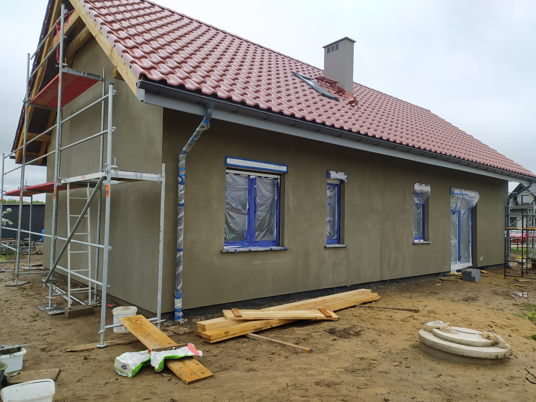 Dom jednorodzinny w trakcie nakładania tynku zewnętrznego, okna zabezpieczone folią, widoczne rusztowania i materiały budowlane, dach pokryty czerwoną dachówką ceramiczną.
