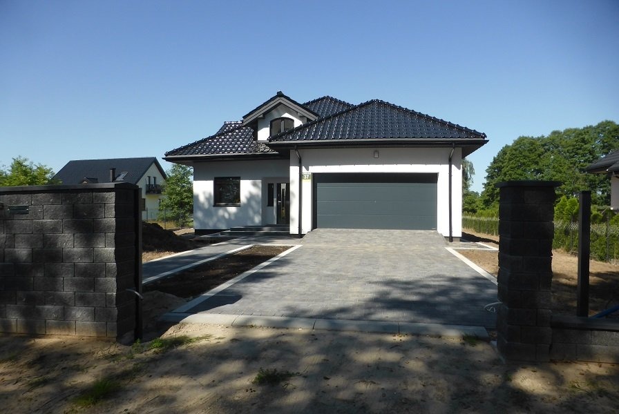 Nowoczesny dom jednorodzinny z szarą bramą garażową i brukowanym podjazdem, widok z poziomu gruntu, z elementami ogrodzenia z szarej kostki.