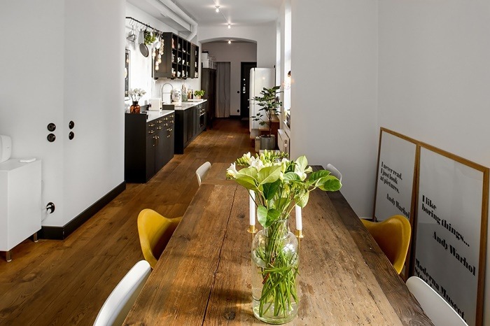 Wnętrze mieszkania z perspektywy stołu z bukietem kwiatów, widok na kuchnię z ciemnymi szafkami i drewnianą podłogą, plakaty Andy'ego Warhola oparte o ścianę.