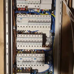 Rozdzielnica elektryczna w obudowie z widocznymi bezpiecznikami, przewodami w kolorach niebieskim, żółto-zielonym i brązowym oraz listwami zaciskowymi, zamontowana na drewnianej ścianie.