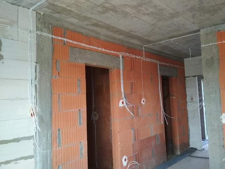 Surowy stan wnętrza domu jednorodzinnego w trakcie budowy, widoczne przewody elektryczne prowadzone po ścianach z czerwonej cegły, przygotowane otwory pod gniazdka, surowy betonowy sufit.