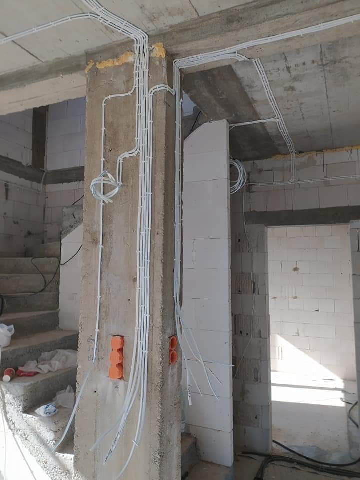 Surowe wnętrze domu jednorodzinnego w trakcie prac instalacyjnych: widoczne kable elektryczne prowadzone natynkowo w białych rurkach PCV, pomarańczowe puszki instalacyjne w betonowym słupie,...