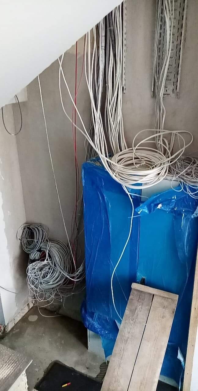 Surowe wnętrze w trakcie prac elektrycznych: wiązki kabli zwisają ze ściany i sufitu, częściowo ułożone w kłęby na podłodze, a szafa elektryczna zabezpieczona niebieską folią czeka na podłączenie.