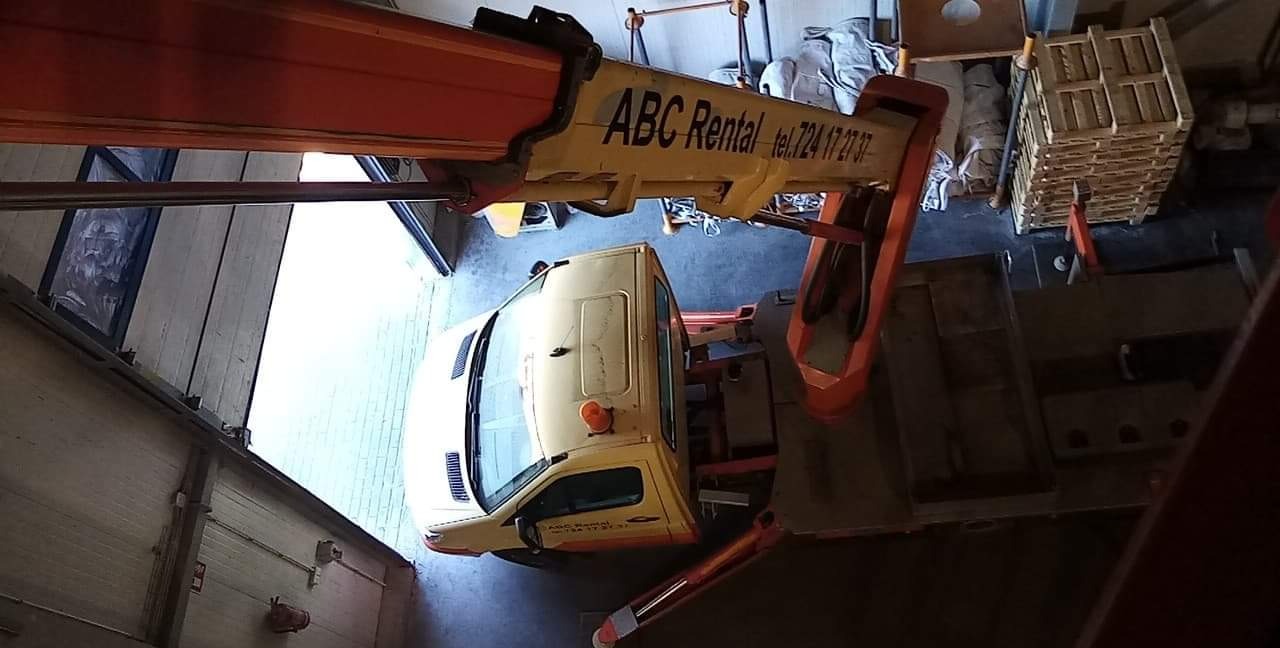 Żółta ciężarówka z dźwigiem firmy ABC Rental, widok z góry, w tle palety i elementy konstrukcyjne budynku.