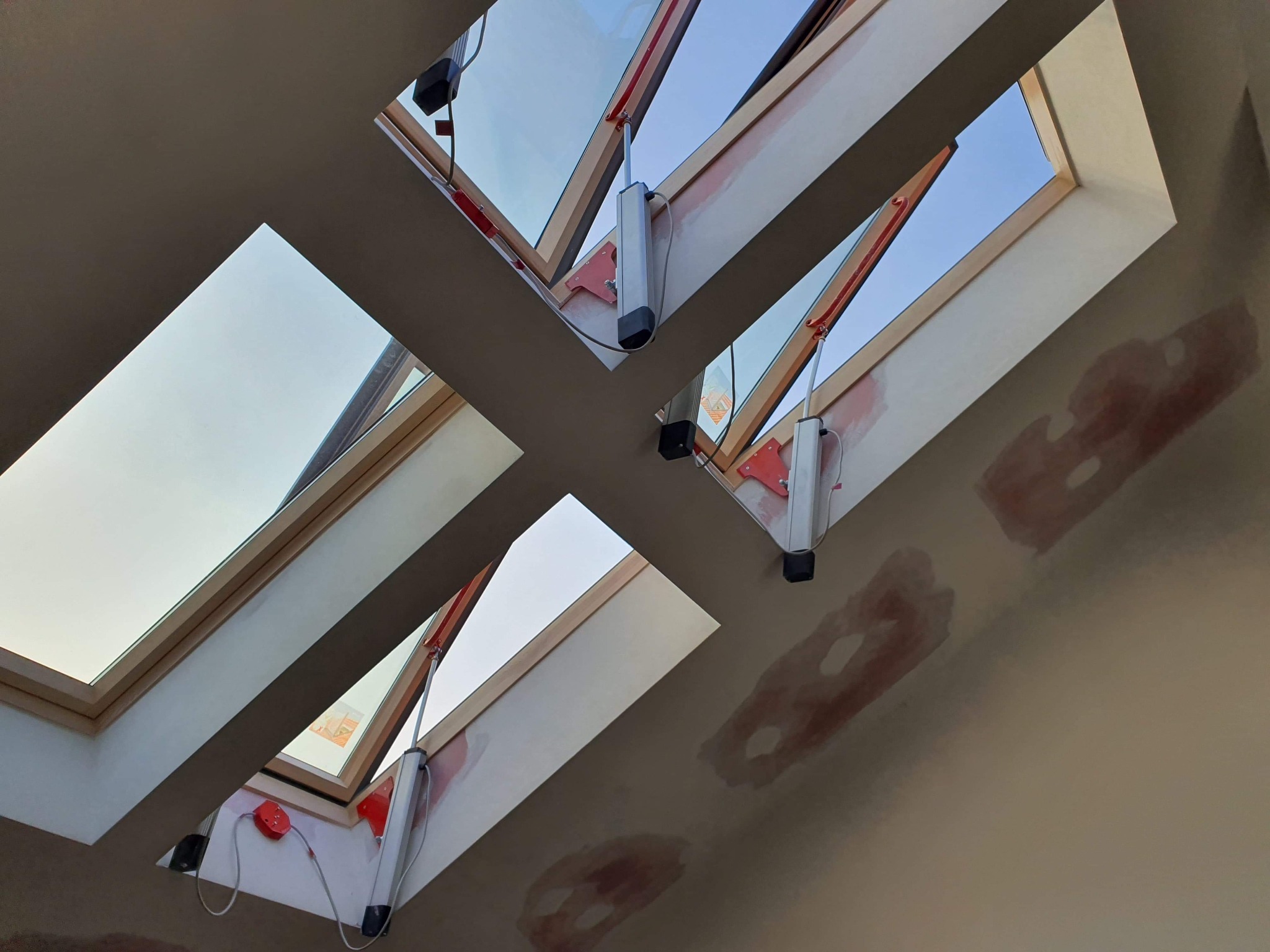 Dwa okna dachowe z zamontowanymi siłownikami elektrycznymi do automatycznego otwierania, widoczne ślady poprawek malarskich na suficie.