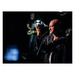 Ck Studio | Photography  - Wokalista z brodą w czarnej marynarce śpiewa do mikrofonu na scenie, w tle muzyk z saksofonem i ciemne oświetlenie.