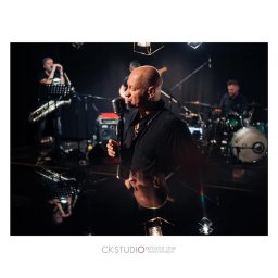 Ck Studio | Photography  - Wokalista z mikrofonem śpiewa na scenie z zespołem w tle, widoczne odbicie w podłodze, oświetlenie sceniczne.