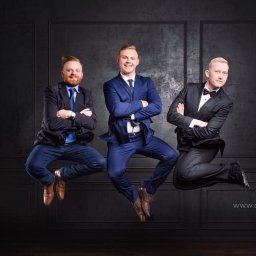 Ck Studio | Photography  - Trzech elegancko ubranych mężczyzn w garniturach, pozujących w skoku na tle ciemnej ściany z panelami, z widocznym logo studia fotograficznego w prawym dolnym rogu.
