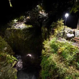 Nocny widok na wodospad w głębokim wąwozie, oświetlony latarniami i reflektorami, z widoczną ścieżką i barierkami.