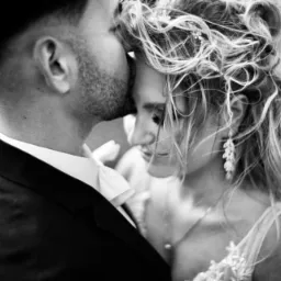 Czarno-białe zdjęcie pary młodej: mężczyzna w garniturze całuje kobietę w czoło, widoczne delikatne kolczyki i upięte włosy panny młodej.