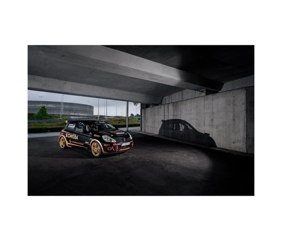 Czarny samochód rajdowy z żółtymi felgami i naklejkami sponsorskimi, sfotografowany pod betonowym wiaduktem, z cieniem samochodu widocznym na ścianie.