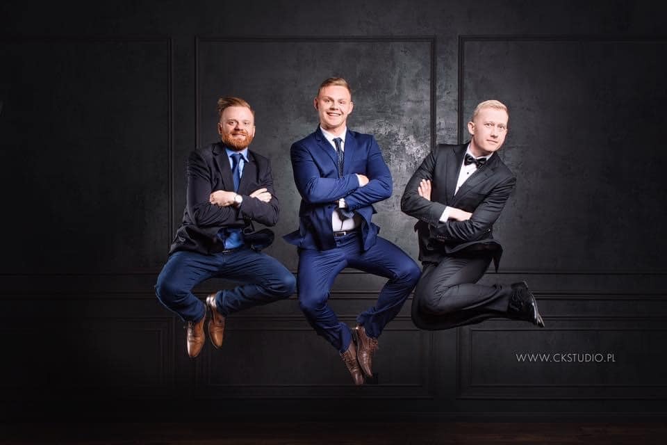 Trzech elegancko ubranych mężczyzn w garniturach, pozujących w skoku na tle ciemnej ściany z panelami, z widocznym logo studia fotograficznego w prawym dolnym rogu.