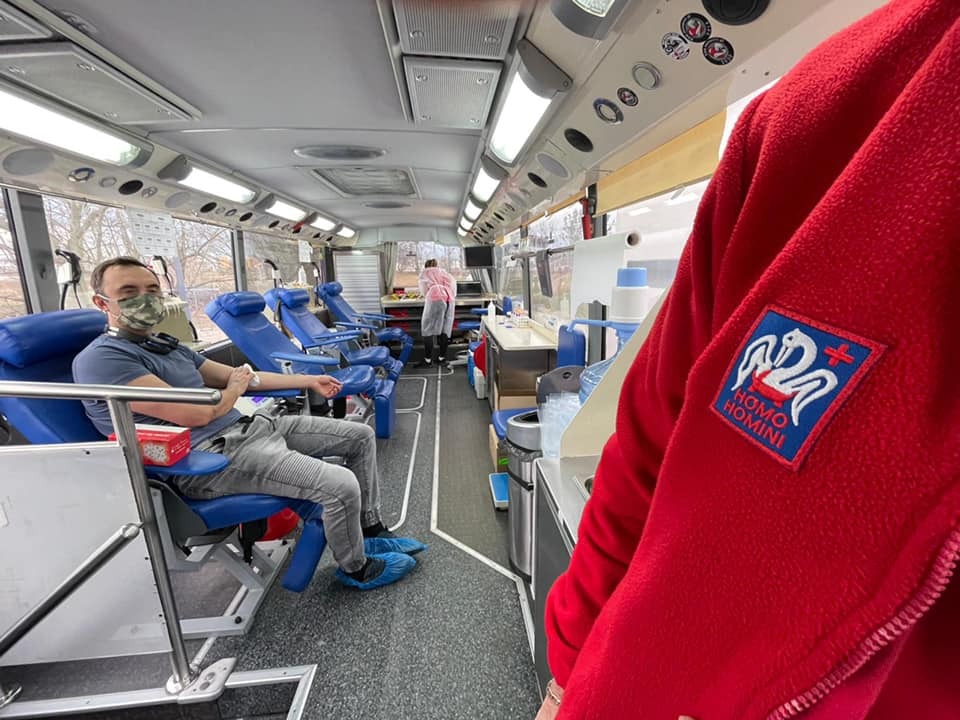 Wnętrze mobilnego punktu poboru krwi: mężczyzna w maseczce siedzi na niebieskim fotelu podczas donacji, w tle widoczny personel medyczny i puste fotele, na pierwszym planie fragment czerwonej bluzy...