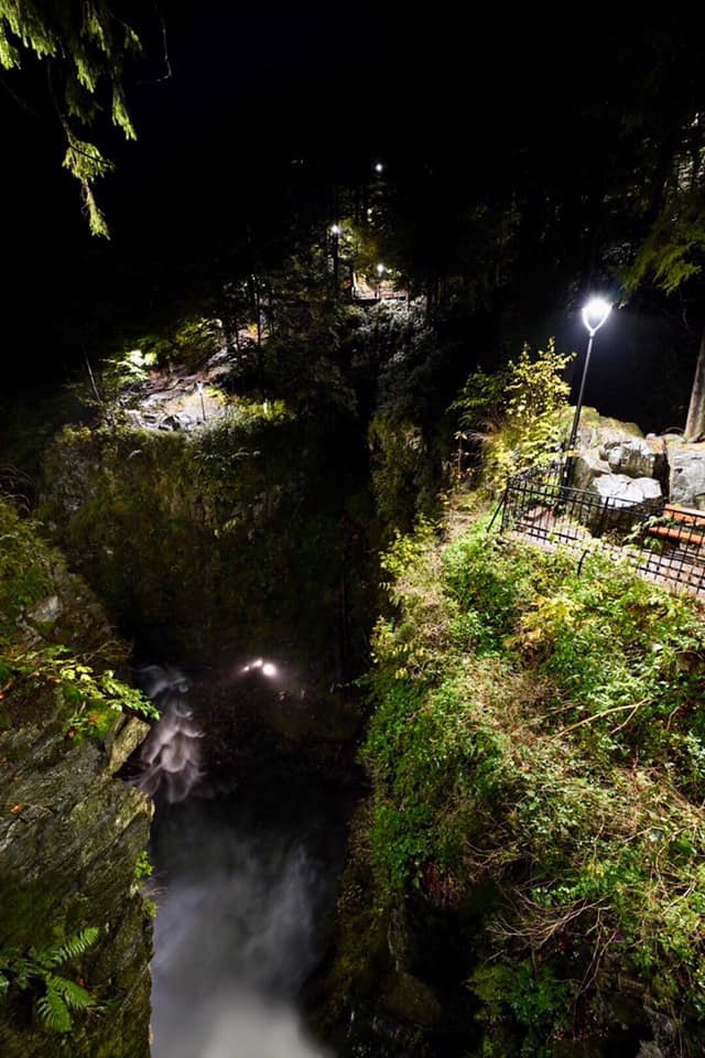 Nocny widok na wodospad w głębokim wąwozie, oświetlony latarniami i reflektorami, z widoczną ścieżką i barierkami.