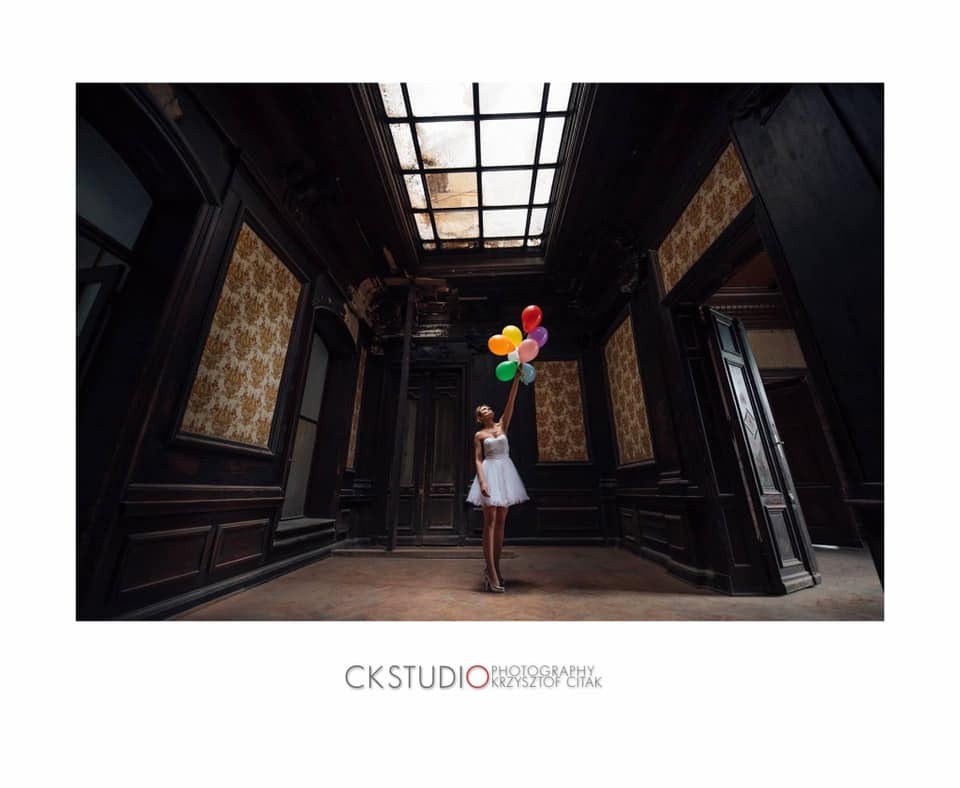 Kobieta w białej sukience trzymająca kolorowe balony w opuszczonym, ciemnym pokoju z oknem w suficie, wzorzyste tapety na ścianach.