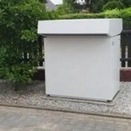 Biała, prostopadłościenna obudowa urządzenia na zewnątrz, otoczona szarym żwirem, z zielenią w tle i fragmentem ogrodzenia.