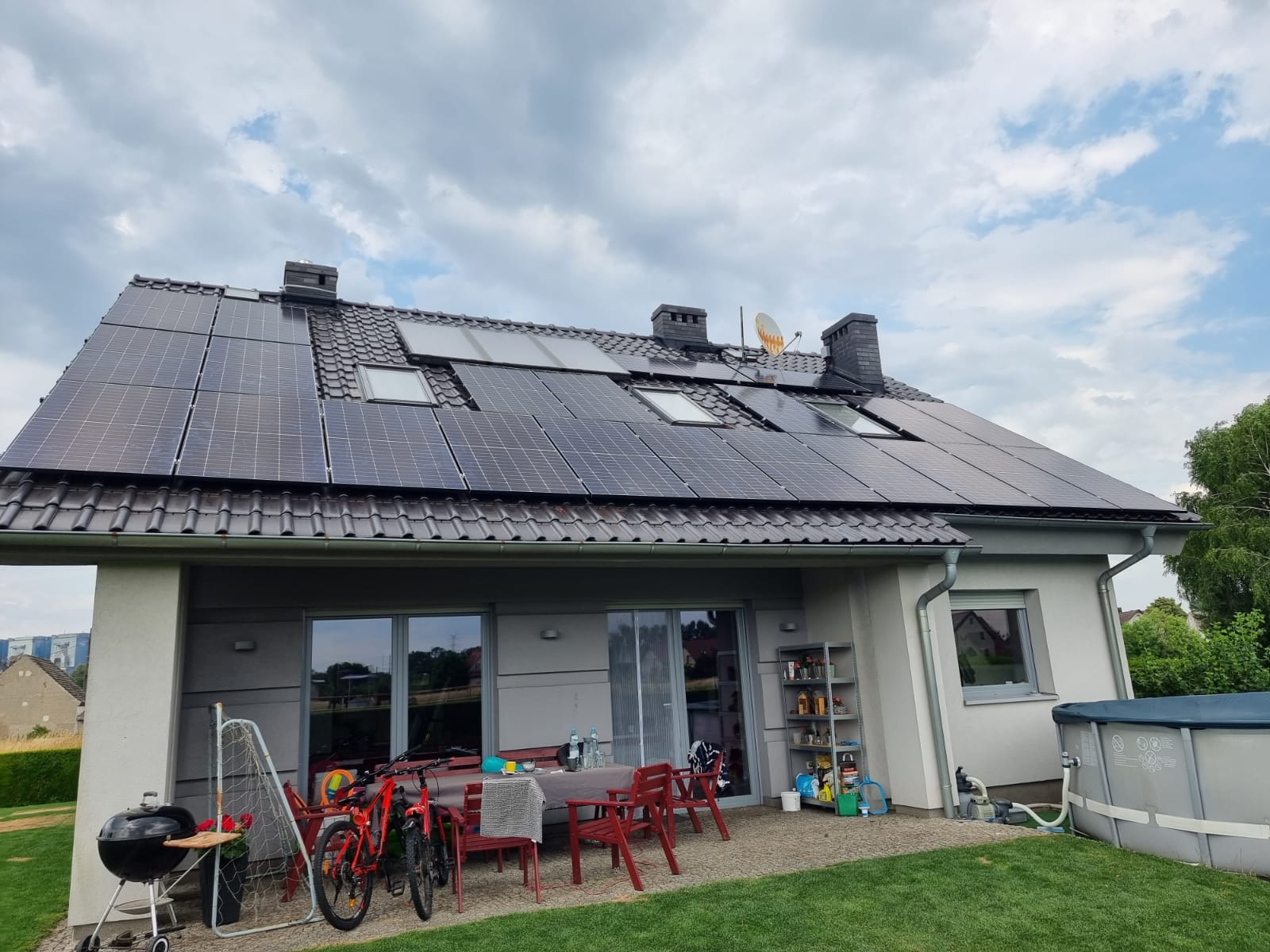 Dom jednorodzinny z panelami słonecznymi na dachu, widoczne okna dachowe, antena satelitarna, taras z grillem i rowerami, pochmurne niebo.