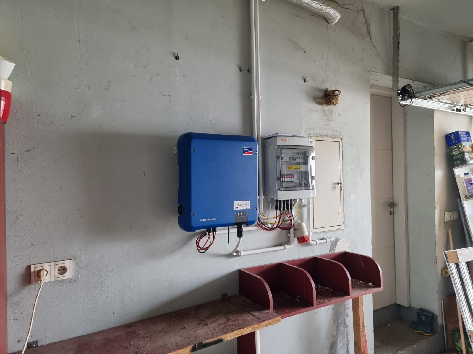 Niebieski falownik SMA i skrzynka elektryczna z zabezpieczeniami zamontowane na szarej, popękanej ścianie w garażu, widoczne przewody i rury instalacyjne.