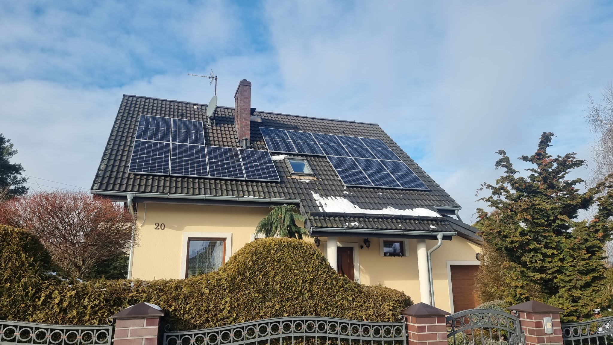 Żółty dom jednorodzinny z ciemną dachówką, na której zamontowane są panele słoneczne; widoczny komin, antena satelitarna i fragmenty śniegu na dachu; ogród z ozdobnymi krzewami i metalowym...