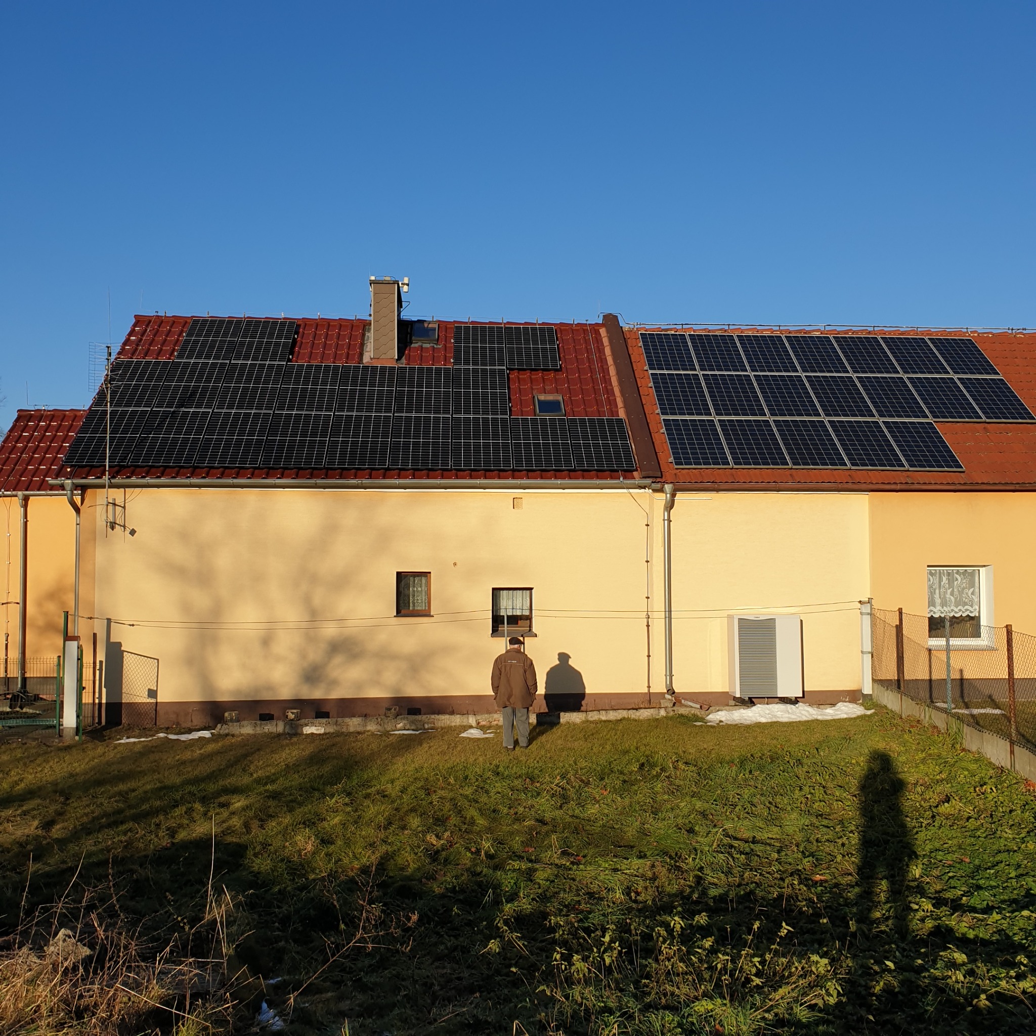 Dom jednorodzinny z panelami fotowoltaicznymi na czerwonym dachu, widoczny również klimatyzator na ścianie i osoba na trawniku rzucająca cień.