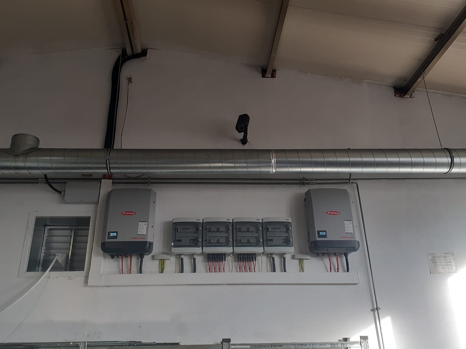 Dwa inwertery Fronius symo i cztery skrzynki zabezpieczeń zamontowane na białej ścianie pod rurą wentylacyjną, widoczna instalacja elektryczna.