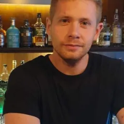 Portret młodego mężczyzny w czarnej koszulce na tle baru z podświetlanymi półkami i butelkami alkoholu.