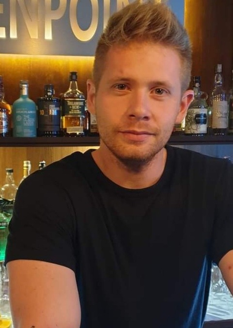 Portret młodego mężczyzny w czarnej koszulce na tle baru z podświetlanymi półkami i butelkami alkoholu.