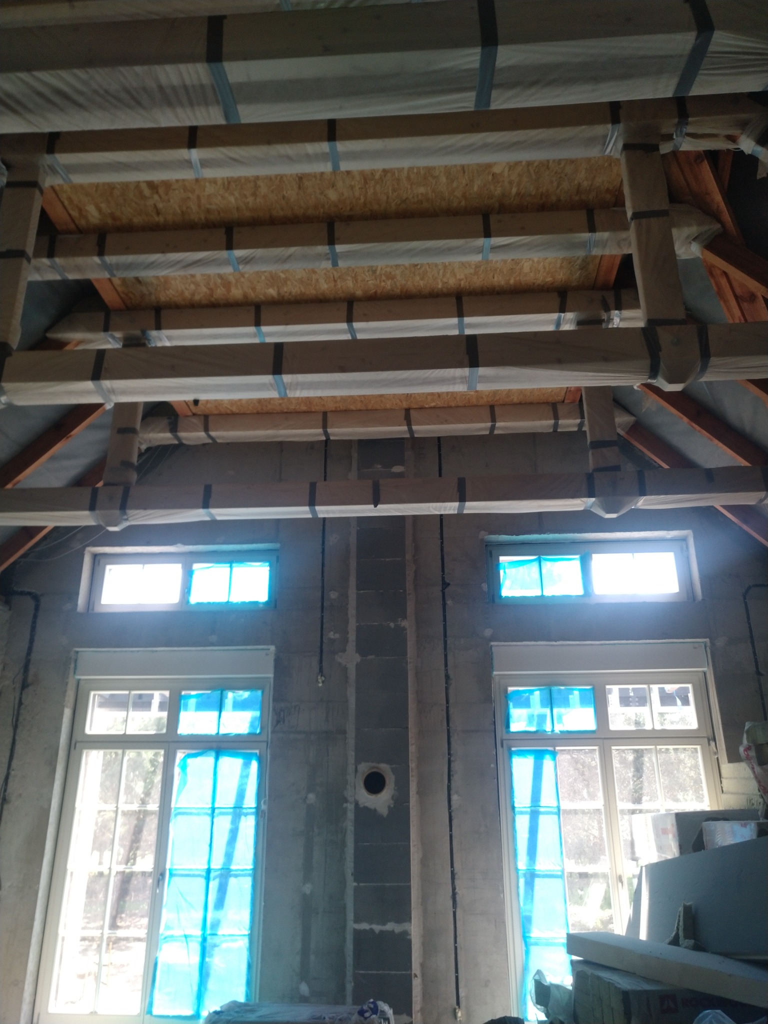 Poddasze w trakcie prac: drewniana konstrukcja dachu zabezpieczona folią i taśmą, okna zakryte niebieską folią, surowe ściany z betonu komórkowego.