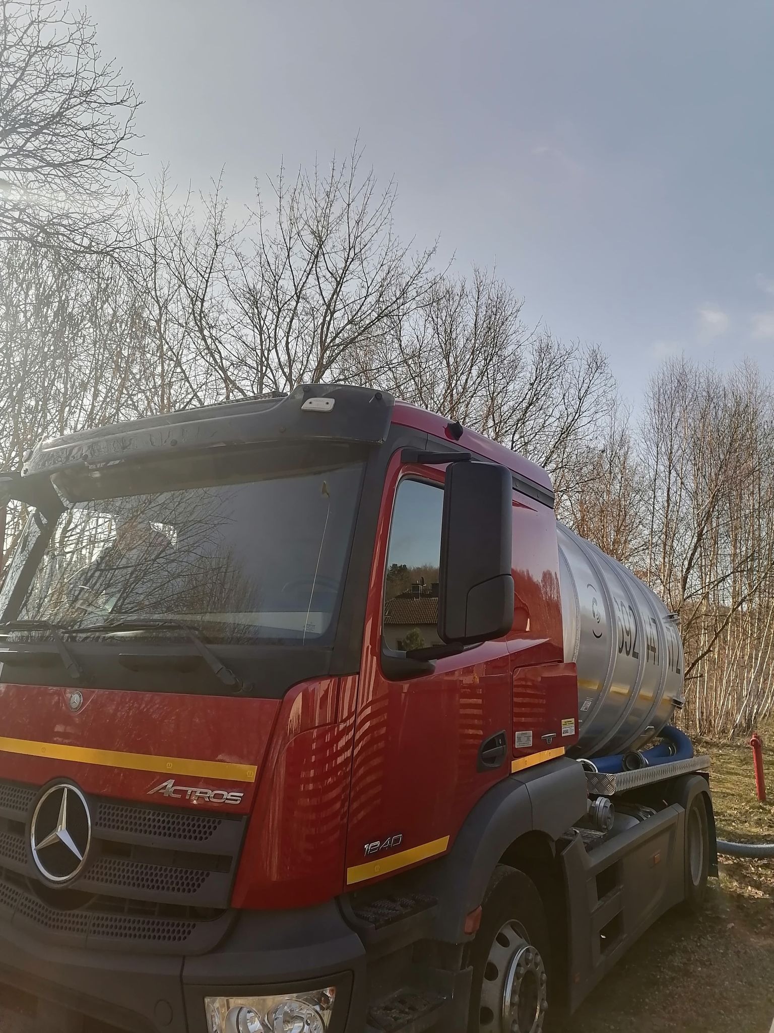 Czerwony samochód ciężarowy Mercedes Actros z cysterną, zaparkowany na zewnątrz z widocznym wężem, prawdopodobnie podczas opróżniania zbiornika, na tle drzew bez liści i częściowo zachmurzonego nieba.