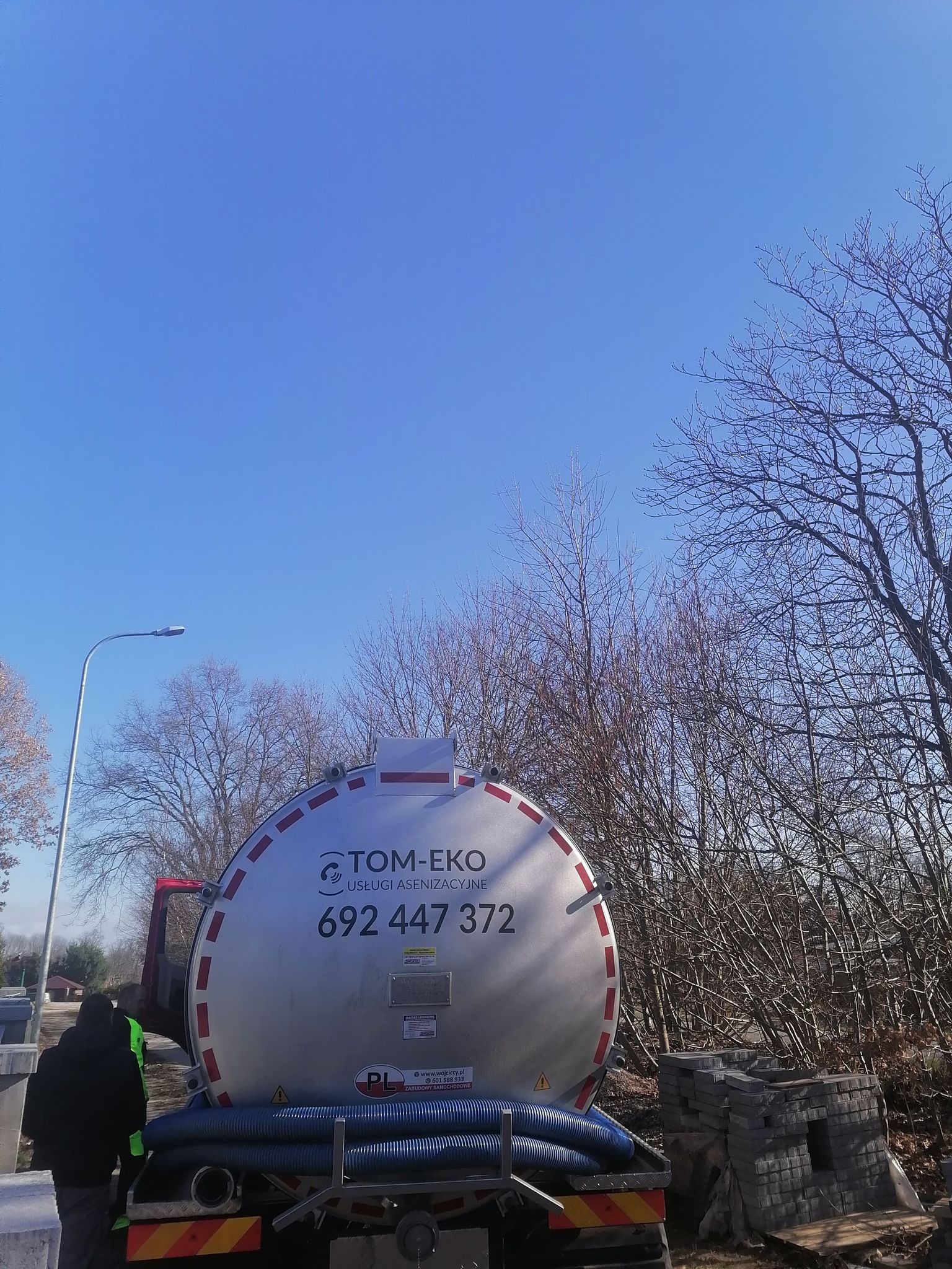Cysterna asenizacyjna firmy TOM-EKO z numerem telefonu 692 447 372 na tle błękitnego nieba i drzew bez liści, obok ułożone kostki brukowe.