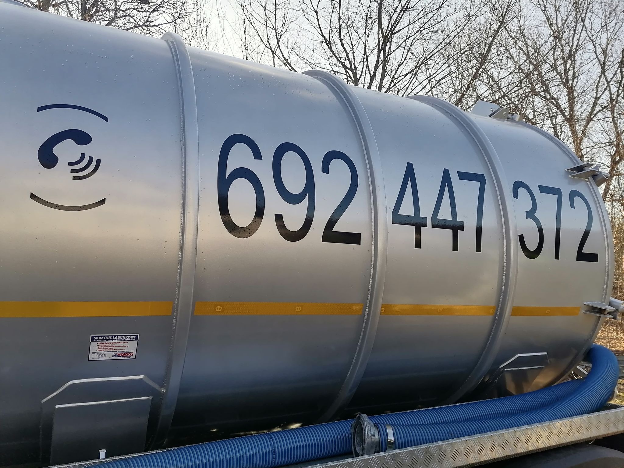 Cysterna asenizacyjna z niebieskim wężem, oznaczona numerem telefonu i symbolem słuchawki, na tle drzew bez liści.