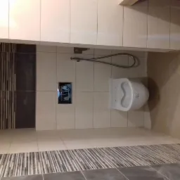 Nowoczesna toaleta podwieszana w łazience z beżowymi płytkami, akcentami ciemnoszarych płytek dekoracyjnych i chromowaną słuchawką prysznicową.