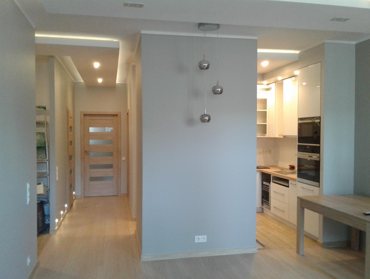Wnętrze mieszkania po remoncie: widok na korytarz z drewnianymi drzwiami i oświetleniem podłogowym oraz otwartą kuchnię z białymi szafkami i sprzętem AGD, całość utrzymana w jasnej kolorystyce...