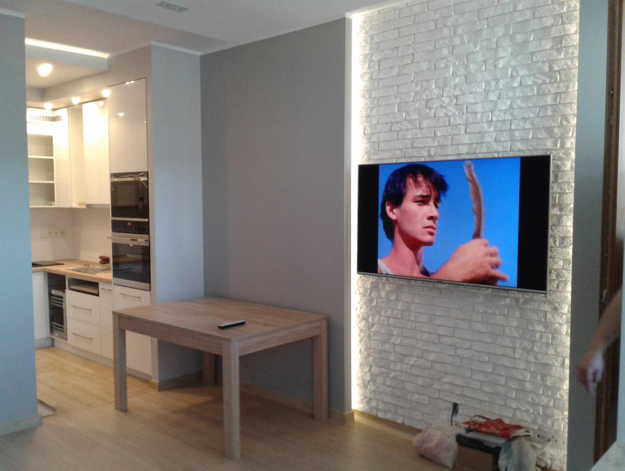 Nowoczesne wnętrze: kuchnia z białymi szafkami, drewniany stół, ściana z białej cegły z podświetleniem LED i zawieszonym telewizorem wyświetlającym film, jasna podłoga.