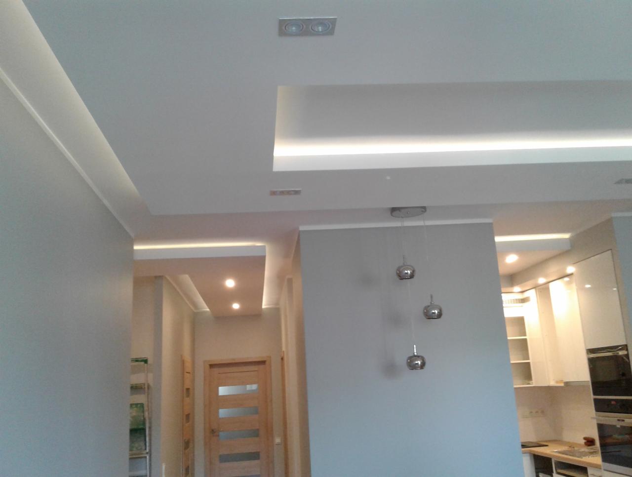 Nowoczesne oświetlenie sufitowe LED w salonie z widokiem na kuchnię, srebrne lampy wiszące, jasne ściany i drzwi z przeszkleniami.