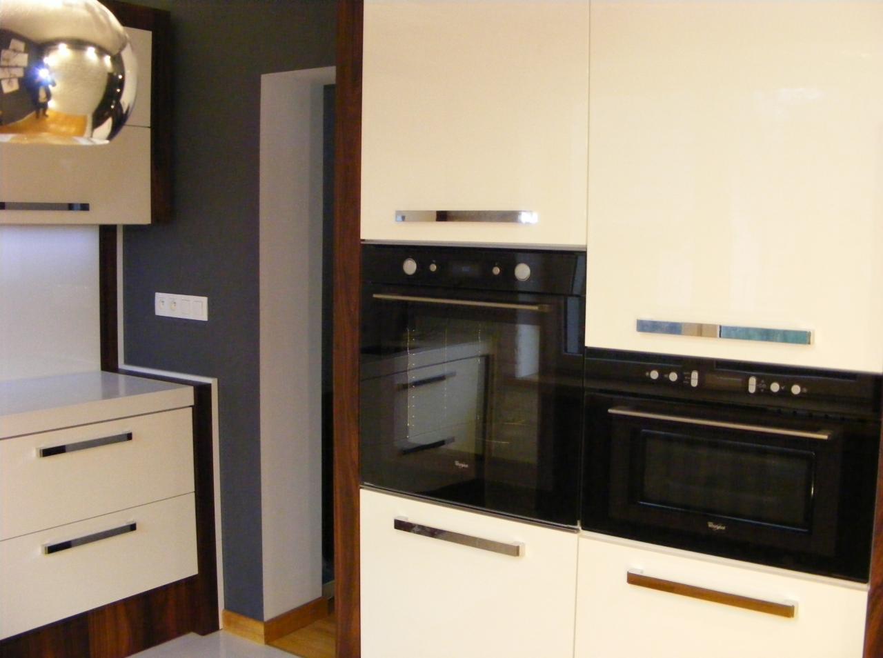 Fragment nowoczesnej kuchni z kremowymi szafkami i wbudowanymi czarnymi piekarnikami Whirlpool, widoczne chromowane uchwyty i odbicie w kulistej lampie sufitowej.