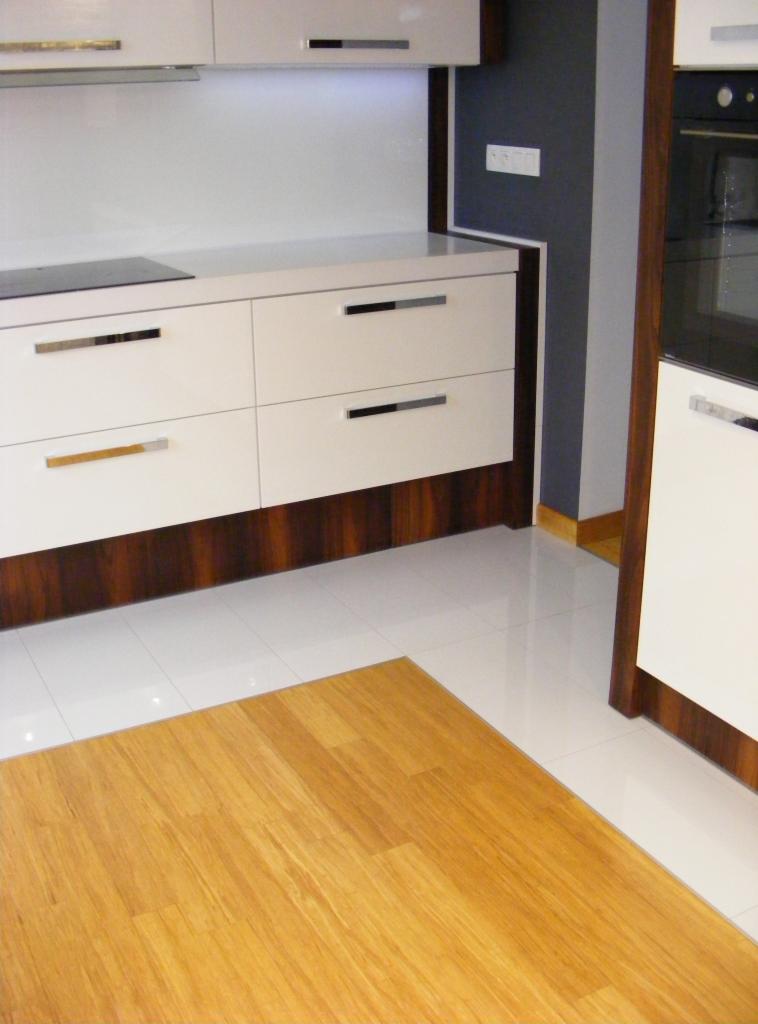 Nowoczesna kuchnia z białymi, lakierowanymi szafkami i ciemnobrązowym fornirem u dołu, białe płytki podłogowe przechodzące w drewnianą podłogę, widoczny fragment zabudowy piekarnika i płyta...