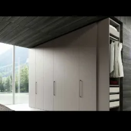 Zamknięta szafa z przesuwnymi drzwiami w kolorze taupe, z otwartą boczną sekcją ukazującą wieszaki z ubraniami i półki z tekstyliami, na tle panoramicznego okna z widokiem na las.