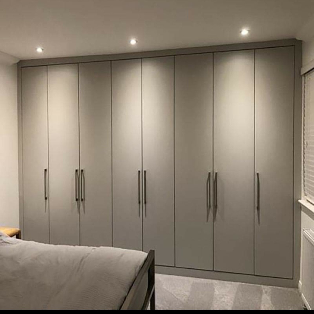 Duża, szara szafa wnękowa z długimi, pionowymi uchwytami, zabudowana od podłogi do sufitu w sypialni z łóżkiem i oświetleniem punktowym.