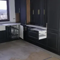 Ciemna zabudowa kuchenna z otwartą szufladą i cargo, złote uchwyty, podłoga zabezpieczona kartonem, widok na krajobraz za oknem.