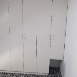 M N M Tomasz Dąbrowski - Biała szafa wnękowa z czterema drzwiami, z minimalistycznymi uchwytami, dopasowana do skosu sufitu, na podłodze wzorzyste płytki w odcieniach szarości.