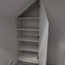 M N M Tomasz Dąbrowski - Biała szafa wnękowa z otwartymi drzwiami, dopasowana do skosu dachu, na podłodze wzorzyste płytki. Widoczne półki wewnątrz szafy.