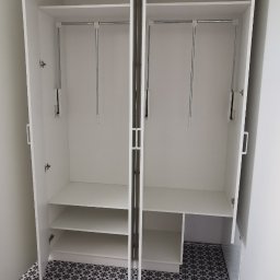 M N M Tomasz Dąbrowski - Biała szafa wnękowa z otwartymi drzwiami, widoczne półki i drążki na ubrania. Na podłodze wzorzyste płytki. Minimalistyczny design.