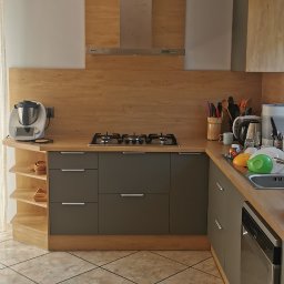 M N M Tomasz Dąbrowski - Nowoczesna kuchnia w odcieniach szarości i drewna z blatem roboczym, płytą gazową i okapem. Widoczny sprzęt AGD i akcesoria kuchenne. Ujęcie z perspektywy podłogi.