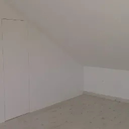 Biała szafa wnękowa z frontami bez uchwytów, wkomponowana w skos strychu, z widocznym fragmentem drzwi wejściowych do pomieszczenia.