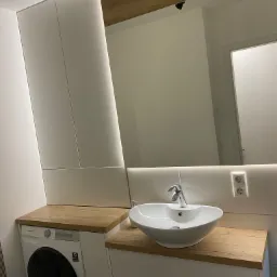 Nowoczesna łazienka z białą szafką pod umywalkę z drewnianym blatem, pralką w zabudowie pod blatem i dużym lustrem z oświetleniem LED.