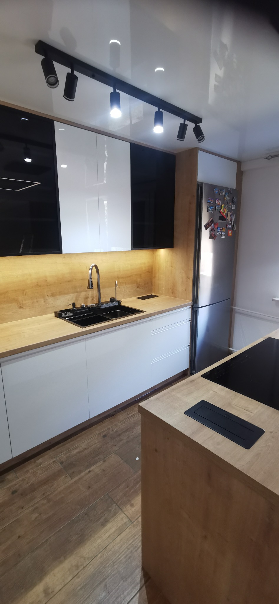Nowoczesna kuchnia z białymi frontami szafek dolnych, czarno-białymi szafkami górnymi, drewnianym blatem i czarnym zlewozmywakiem, oświetlona reflektorami.