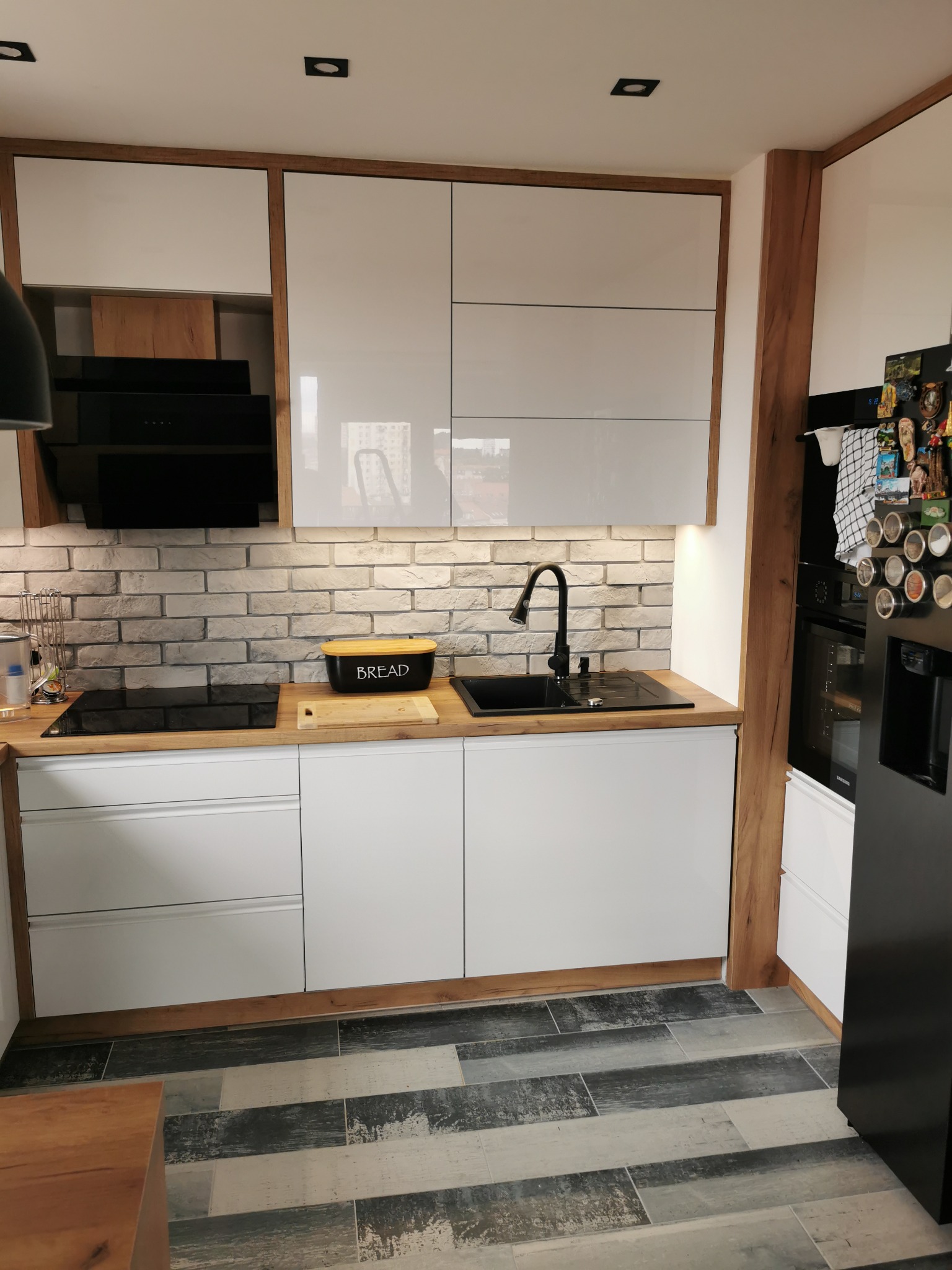 Nowoczesna kuchnia z białymi, lakierowanymi frontami, drewnianymi akcentami, ceglaną ścianą i czarną armaturą, widok na płytę indukcyjną, zlew i piekarnik.